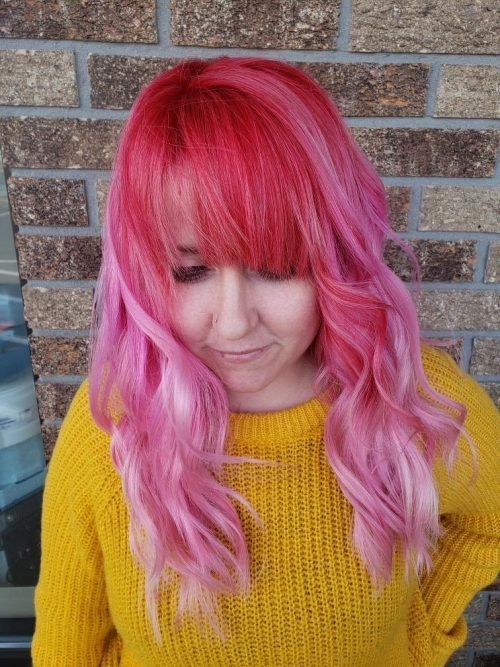 Dani-Winterhart Pink haired woman stands in front of a brick wall outside, wearing a yellow sweater and looking down.